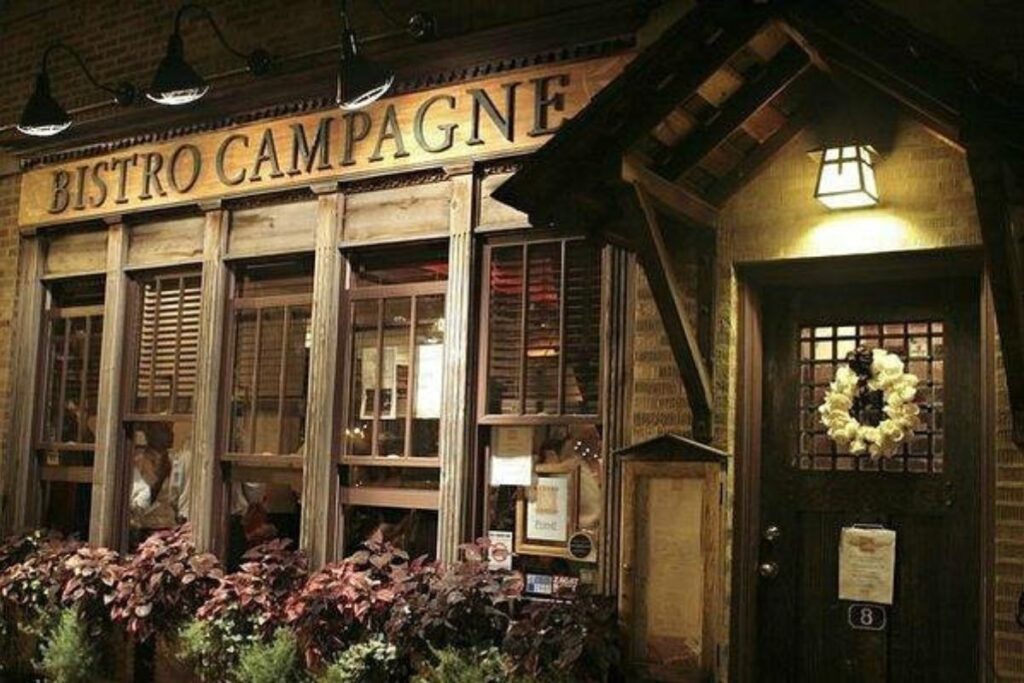 Warm wooden storefront of Bistro Campagne lit at night with a lantern and flower boxes in Lincoln Square, Chicago