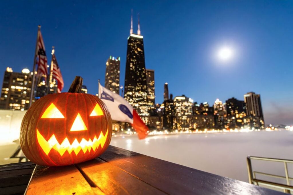 Lit jack-o'-lantern on a railing at night with the Chicago skyline and John Hancock Center behind it