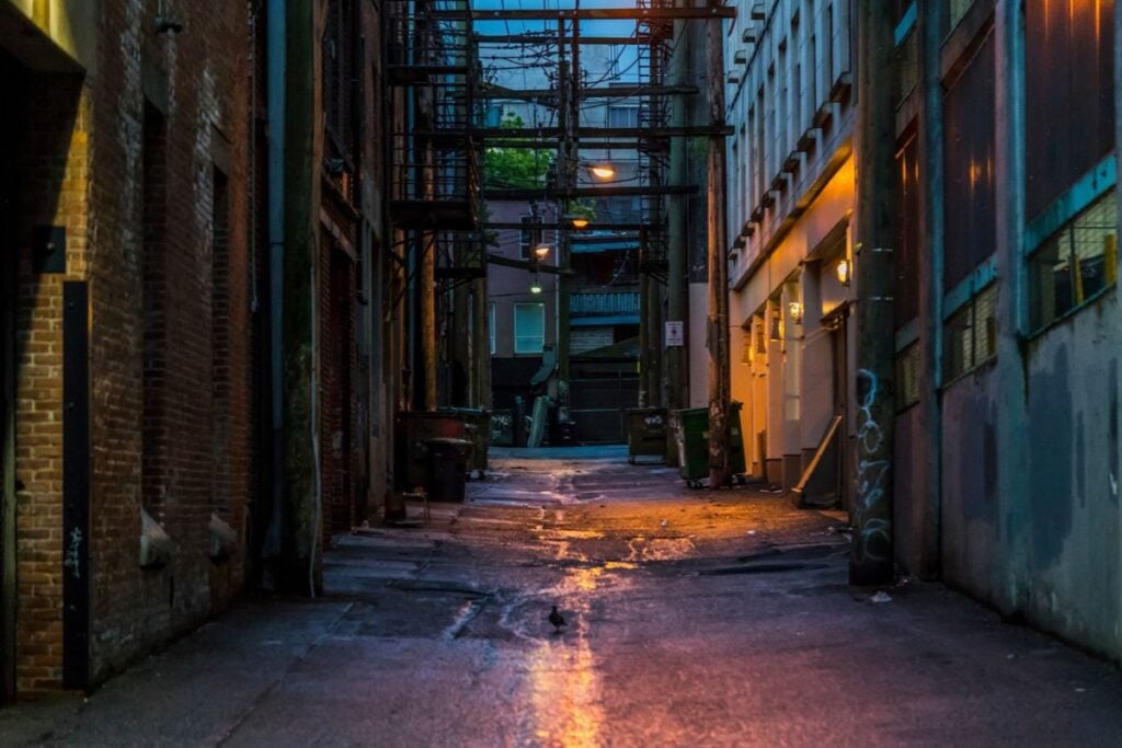 Empty city alley at dusk with wet pavement, fire escapes, and dim lamps glowing on brick walls