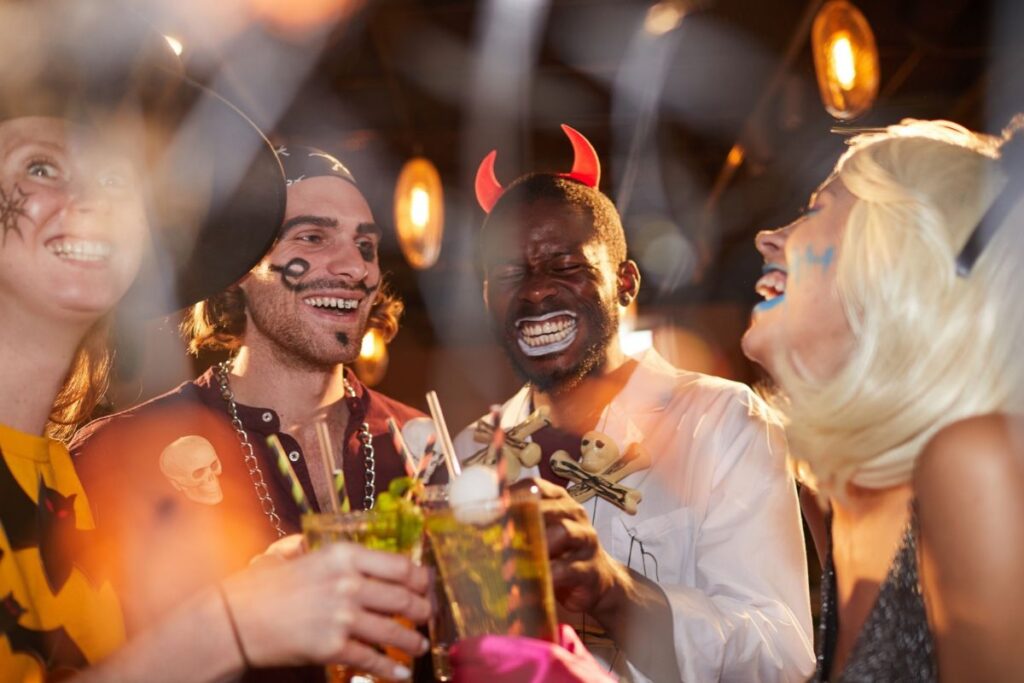 Friends in Halloween costumes, including devil horns and pirate face paint, toasting with cocktails at a bar