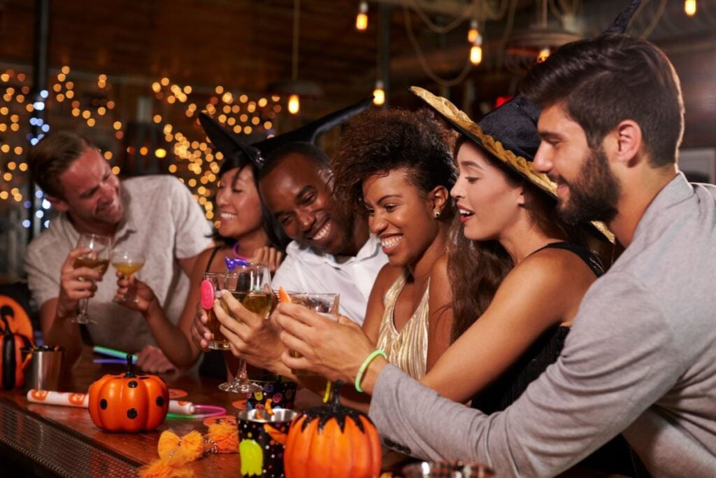 Group of friends in witch hats toasting at a bar decorated with pumpkins and string lights for Halloween